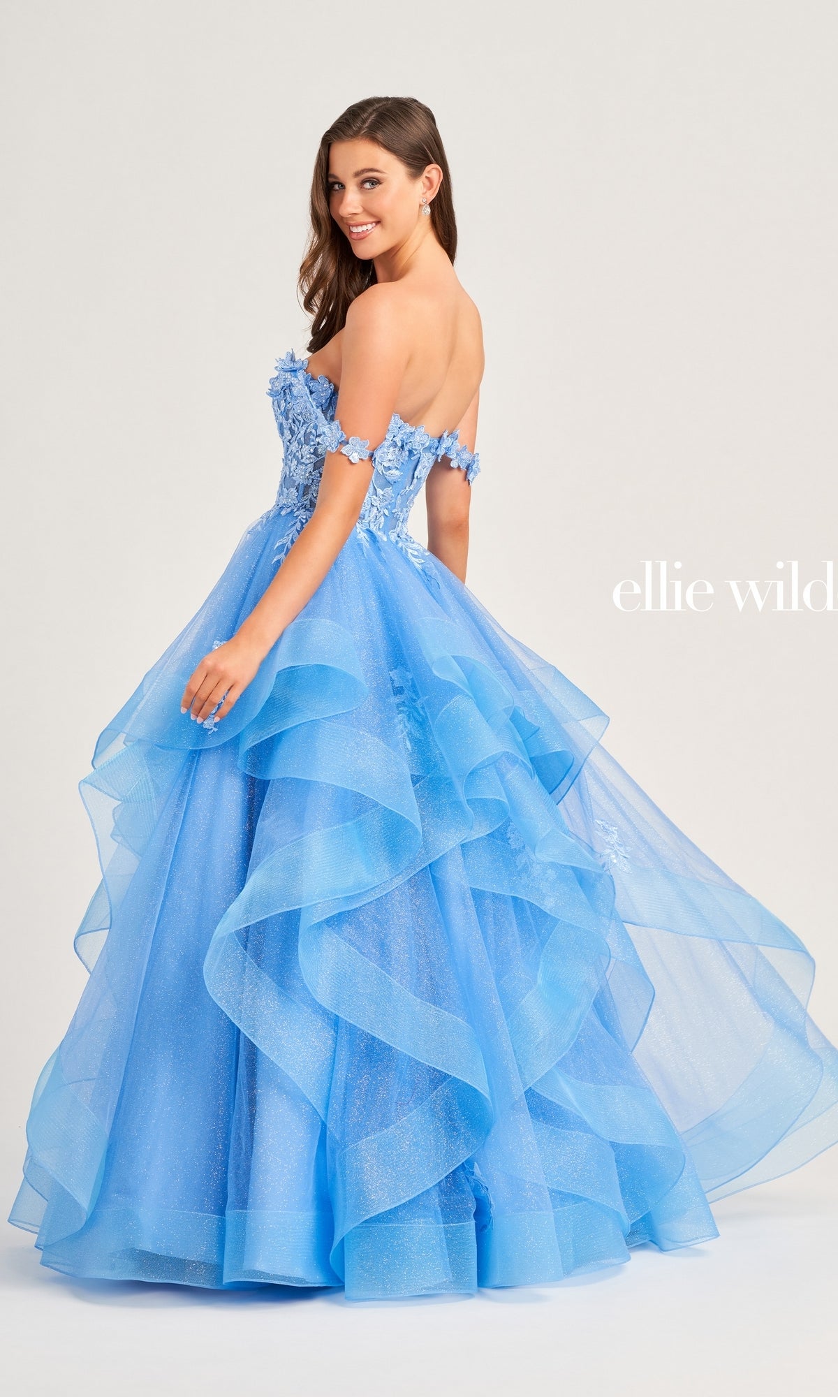 A woman stands smiling over her shoulder in the Ellie Wilde Long Designer Prom Ball Gown EW35084, a strapless, tiered blue glitter tulle dress featuring floral appliqué details.